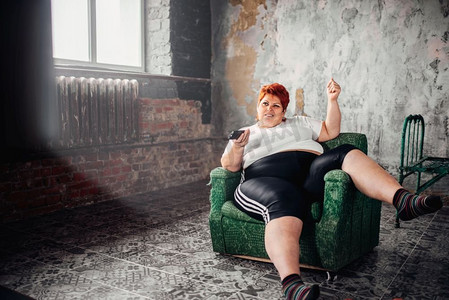 超重的女人坐在椅子上看电视。不健康的生活方式，肥胖。超重的女人坐在椅子上看电视