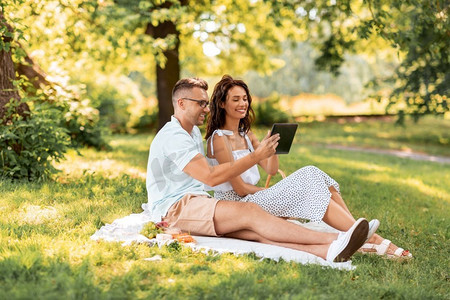 休闲与人的概念快乐的夫妇和平板电脑在夏令营野餐。带着平板电脑在公园野餐的幸福夫妻