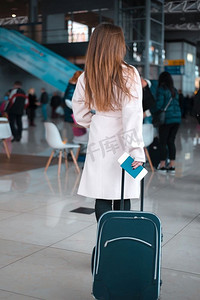 拖着行李箱的人摄影照片_年轻的女旅行者在机场大厅拖着行李。机场咖啡馆背景。在机场大厅散步的旅客。