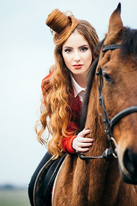 骑师摄影照片_红发骑师女孩，穿着红色羊毛衫和黑色高靴，牵着马散步