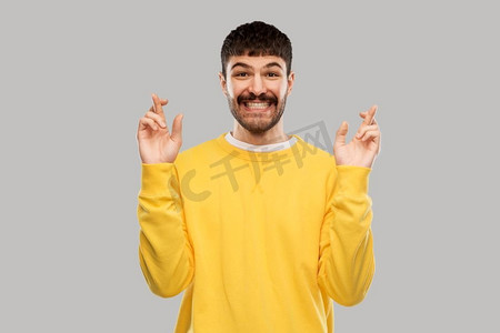 黄色手指手势摄影照片_手势、运气和人的概念—微笑的年轻人交叉手指在灰色背景的黄色运动衫。手指交叉的微笑男人