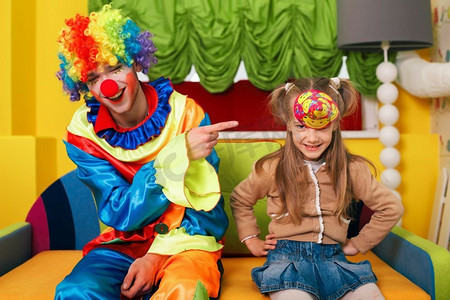 小丑摄影照片_小女孩戴上了帽子，小丑嘲笑她。背景是幼儿园。