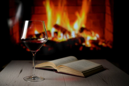 干杯红酒摄影照片_背景中干杯红酒,打开的书和壁炉台灯火堆