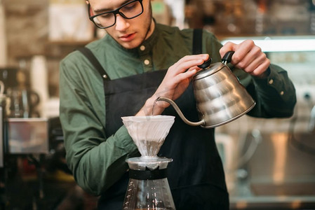 穿着黑围裙、戴眼镜的服务员在吧台上的咖啡壶里倒热水。服务员把热开水倒进咖啡壶里
