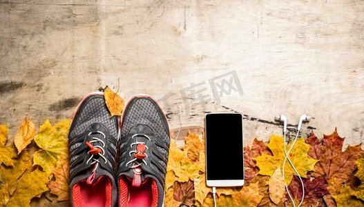 运动鞋在秋天的叶子，手机和耳机。在木制背景上。运动鞋在秋天的叶子，手机和耳机。