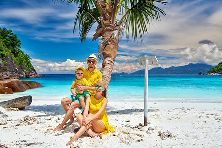 父亲马摄影照片_一家人在美丽的小安塞海滩上，年轻夫妇和三岁的蹒跚学步的男孩坐在棕榈树上。马赫，塞舌尔的暑假。