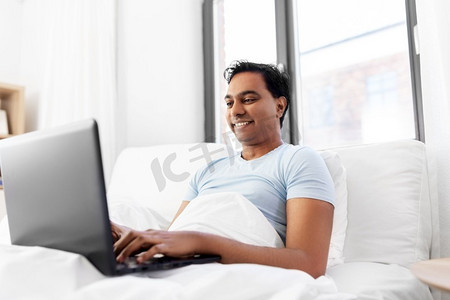 人，技术和休息概念—有膝上型计算机的愉快的微笑印度人躺在床上在家快乐的印度人与笔记本电脑在床上在家里
