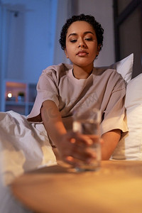 睡前喝水摄影照片_人，就寝时间和口渴概念—非洲裔美国妇女在床上喝水在晚上。非裔美国人妇女在晚上喝水