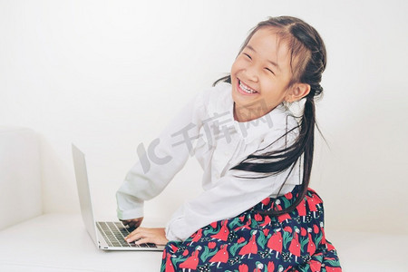 坐在白色沙发上的使用膝上型计算机的小快乐的孩子。童年生活方式使用膝上型计算机的小孩在白色沙发上。