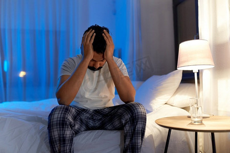 睡眠问题、就寝时间和失眠概念-压力很大的印度男人晚上在家坐在床上。压力很大的印度男子晚上坐在床上