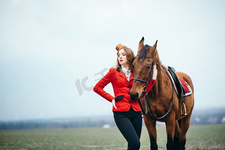 红发骑师女孩，穿着红色羊毛衫和黑色高靴，牵着马散步