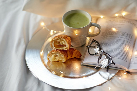 早晨，卫生间和早餐概念--牛角面包、一杯抹茶茶书和家里床上的玻璃杯。床上有牛角面包、抹茶、书和玻璃杯