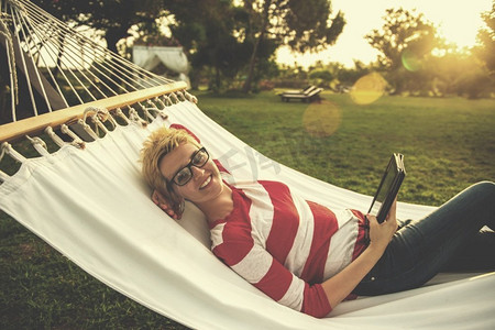 一个年轻女子使用平板电脑，而放松在吊床在宁静的花园在假期