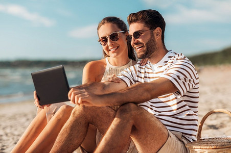休闲和人概念—快乐夫妇与平板电脑电脑和野餐篮在夏季海滩。快乐的夫妇与平板电脑在野餐在海滩