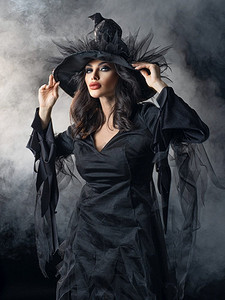 万圣节女巫摄影照片_美丽的女人在万圣节女巫服装礼服和帽子在雾。女人在万圣节女巫服装