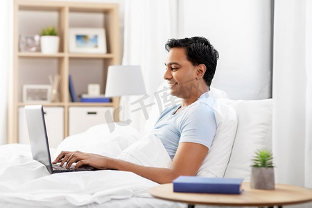 人，就寝时间和休息概念—有膝上型计算机的愉快的微笑印度人躺在床上在家快乐的印度人与笔记本电脑在床上在家里