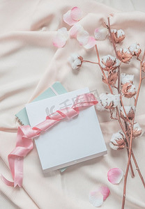 美观的生活方式，笔记本，白色盒子和粉色丝带，米色面料背景，棉枝和粉色花瓣。俯视图。美