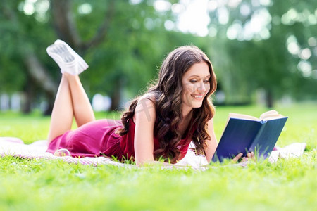 休闲和人的概念—快乐微笑的年轻妇女读书在夏季公园。快乐的微笑的女人读书在夏天公园