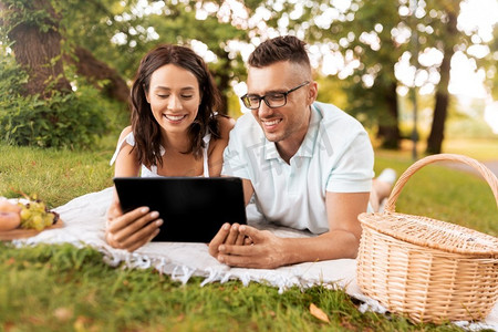 休闲与人概念—快乐夫妇与平板电脑电脑在夏季公园野餐。愉快的夫妇与平板电脑在野餐在公园
