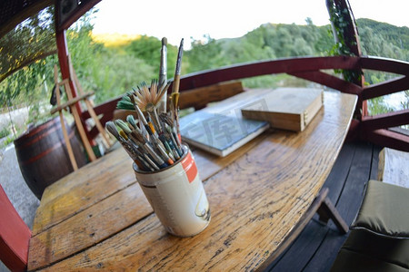 画笔刷背景摄影照片_画家在花园背景的木桌上作画时，画笔刷在罐子里的照片