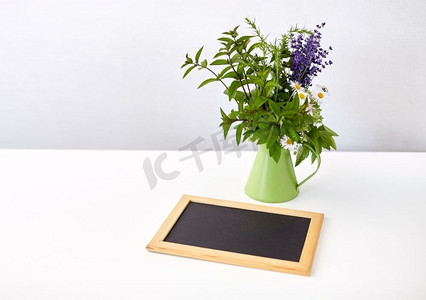 桌子上的罐子摄影照片_园艺，植物和有机概念-绿色罐子里的一束草药和鲜花，桌子上有黑板。一串草药和鲜花配上黑板