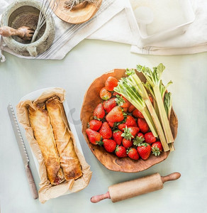 家常菜摄影照片_草莓大黄包装蛋糕在厨房的桌子背景与碗与配料。俯视图。用季节性产品进行夏季烘焙。家常菜