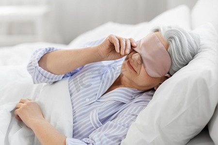 老年人和人的概念—高级妇女与眼睛睡眠面具在床上在家庭卧室。老年妇女与眼罩在床上在家里