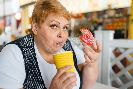 胖女人在快餐店吃甜甜圈，不健康的食物。超重的女性人在桌子上与垃圾晚餐，肥胖问题