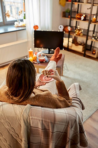 万圣节，假日和休闲概念年轻妇女看电视和吃华夫饼在舒适的家里。女人在万圣节看电视吃华夫饼