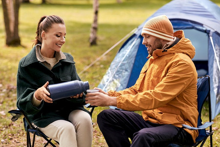 露营、旅游和旅行概念—愉快的夫妇与热水瓶倒茶杯在帐篷营地。快乐的夫妇与热水瓶倒茶在帐篷营地