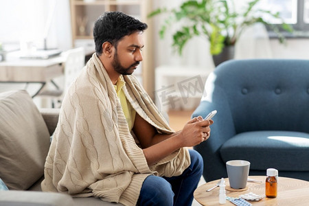 健康，寒冷和人的概念—生病的年轻印度男子毯子与智能手机在家里。生病的年轻人在毯子与智能手机在家里