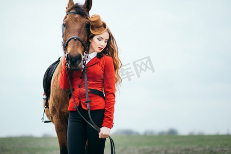 红发骑师女孩，穿着红色羊毛衫和黑色高靴，牵着马散步