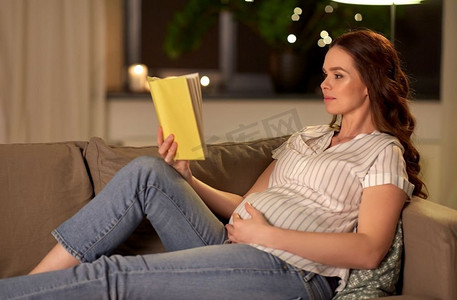 怀孕和人的概念—快乐的微笑孕妇阅读书在家里的沙发上。快乐的微笑孕妇读书在家