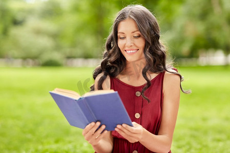 休闲和人的概念—快乐微笑的年轻妇女读书在夏季公园。快乐的微笑的女人读书在公园