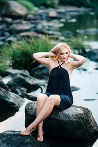 金发女孩在一件黑色的裙子与蓝色的眼睛在河的石头海岸