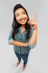 手指指向摄影照片_人物和手势概念-快乐微笑的亚洲年轻女子将手指指向灰色背景。快乐微笑的亚洲女人向上指着手指