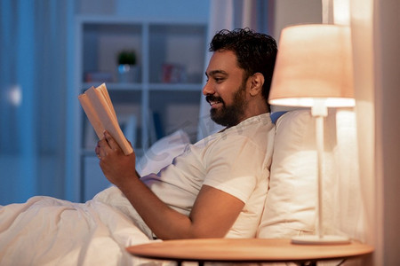 睡觉书摄影照片_人，睡觉时间和休息的概念-快乐的微笑的印度男人晚上在家的床上看书。快乐的印度男人晚上躺在床上看书