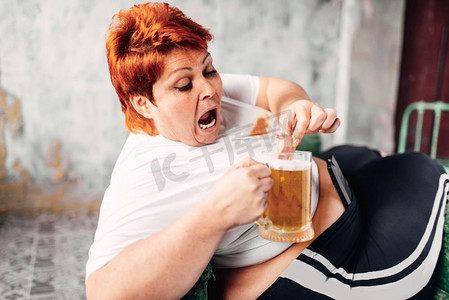 肥胖女性摄影照片_超重的女人喝啤酒，吃三明治，暴食症，肥胖。不健康的生活方式，肥胖的女性。超重的女人喝啤酒，肥胖