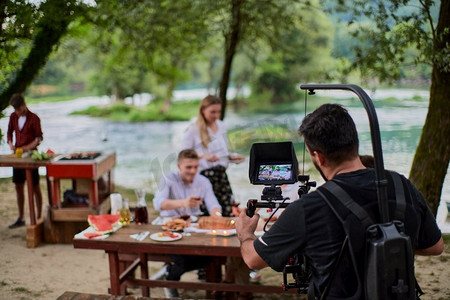 一位配备专业的摄影师正在拍摄一群人在河边吃晚饭，