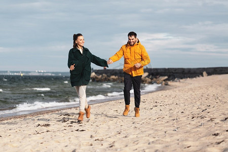 海滩跑步摄影照片_爱情、关系和人的概念-幸福的微笑情侣沿着秋日海滩奔跑，手牵手。一对情侣沿着秋天的海滩跑步