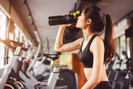 亚洲美女在运动健身训练馆喝蛋白质奶昔或饮用水。美女室内一角
