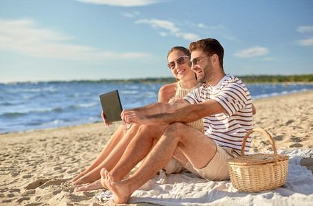 休闲和人概念—快乐夫妇与平板电脑电脑和野餐篮在夏季海滩。快乐的夫妇与平板电脑在野餐在海滩
