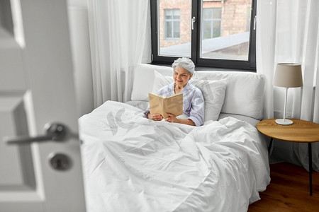 老年，休闲和人的概念—高级妇女阅读书在床上在家里的卧室。高级妇女阅读书在床上在家庭卧室