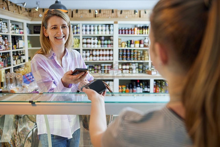 购物app摄影照片_微笑的女顾客Delicatessen食品店用手机非接触式支付购物给销售助理