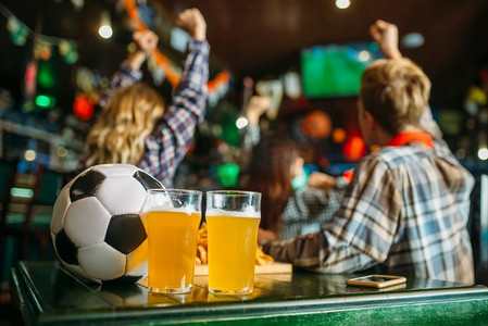 球和啤酒在桌子上在体育酒吧，背景的足球迷电视转播，看游戏概念