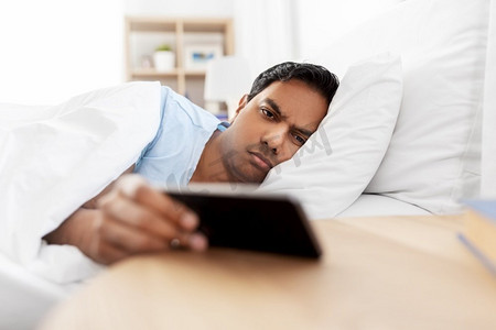 人，睡觉时间和休息的概念-困倦的印度男人躺在床上的智能手机在家里。昏昏欲睡的印度男子躺在床上拿着智能手机