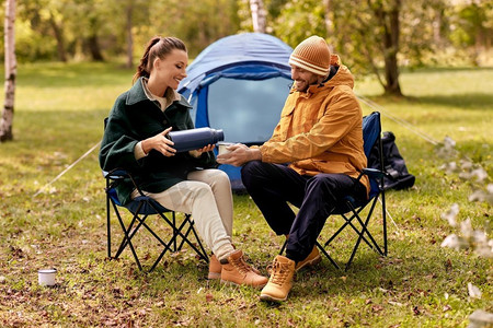 露营、旅游和旅行概念-在帐篷营地，用保温瓶将茶倒到杯子里，这对情侣很幸福。一对幸福的夫妇在帐篷营地用保温瓶倒茶