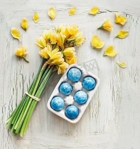 蓝色水仙花摄影照片_蓝色复活节彩蛋夹着黄色水仙花，白色寒酸的小鸡为背景。顶视图