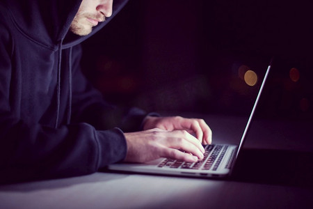 年轻的天才黑客在黑暗的办公室里工作时使用笔记本电脑，晚上背景是大城市的灯光