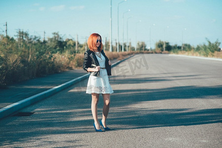 黑色的路摄影照片_一个穿黑夹克、戴蓝眼镜的红头发女孩走在灰色的路上